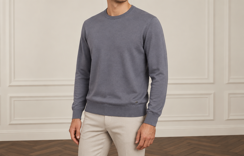 On-model photoshoot (studio)
Show the garment from the image worn by a professional fashion model standing in a professional photo studio with a clean seamless background and soft editorial lighting. Reproduce the garment's exact design, color, fabric and details faithfully. Full-body shot, photorealistic, no text. Additional instructions: Premium men's fashion e-commerce product photography of a viscose knit jumper, matching the established reference composition, pose, body rotation, camera angle, proportions and premium studio aesthetic.
COMPOSITION:
Upper-body men's fashion catalogue photograph.
Show the male model from approximately the LOWER CHIN / NECK AREA down to the UPPER THIGHS.
The FACE must remain completely outside the frame.
No eyes.
No nose.
No full face visible.
Only a small portion of the lower chin and neck may be visible at the very top of the frame.
The complete jumper must be clearly visible:
neckline,
shoulders,
chest,
sleeves,
cuffs,
waist,
and bottom hem.
The jumper must occupy the majority of the image and remain the dominant visual focus.
POSE — ABSOLUTELY CRITICAL:
Use the same established premium catalogue pose.
The model stands with a subtle natural THREE-QUARTER body rotation.
The body must NOT be completely front-facing.
The LEFT SHOULDER is positioned FORWARD and slightly CLOSER TO THE CAMERA.
The RIGHT SHOULDER is positioned slightly FARTHER BACK.
The LEFT SIDE of the torso is therefore slightly closer to the camera.
The rotation must remain subtle, approximately 10–20 degrees.
Do NOT mirror the pose.
Do NOT place the right shoulder forward.
Do NOT make the shoulders symmetrical and frontal.
Do NOT create a strong side profile.
LEFT SHOULDER FORWARD is mandatory.
ARMS AND HANDS:
Both arms hang naturally and STRAIGHT DOWN alongside the body.
Both hands remain naturally positioned beside the outer upper thighs.
Hands are relaxed.
Fingers are naturally relaxed.
No hands in pockets.
No crossed arms.
No bent or raised arms.
No touching the jumper.
No pulling the hem.
No adjusting the sleeves.
No touching the collar.
No fashion gestures.
The pose must look calm, masculine, sophisticated and effortless.
JUMPER — ABSOLUTE PRODUCT PRESERVATION:
Use the EXACT viscose jumper from the supplied source image.
Preserve the exact:
design,
color,
shade,
silhouette,
fit,
proportions,
length,
shoulder width,
chest width,
sleeve length,
sleeve width,
neckline,
collar construction,
cuffs,
bottom hem,
ribbing if present,
knit structure,
stitching,
seams,
logos or details if originally present,
and all original construction details.
Do NOT redesign the jumper.
Do NOT change its color.
Do NOT change its original fit.
Do NOT make it tighter.
Do NOT make it oversized.
Do NOT shorten or lengthen it.
Do NOT change the neckline.
Do NOT change the sleeves.
Do NOT add details that are not present in the source image.
The generated jumper must look like the EXACT SAME physical product as in the source photograph.
VISCOSE FABRIC — CRITICAL:
The jumper is made from premium VISCOSE knit fabric.
The material must clearly retain the characteristic appearance and behavior of high-quality viscose.
The fabric should look:
smooth,
soft,
fine,
refined,
fluid,
supple,
elegant,
and premium.
Viscose must have a naturally smooth surface with a very subtle refined luster.
The sheen must be extremely delicate and realistic.
NEVER make the fabric excessively shiny.
NEVER make it look glossy.
NEVER make it look like satin, silk, polyester or synthetic plastic.
The viscose knit must have a soft, fluid and elegant drape.
The fabric should fall naturally over the torso rather than appearing stiff or rigid.
Show realistic gentle folds around the waist, sides, elbows and sleeves.
The folds should be soft and controlled, reflecting the natural fluidity of viscose.
The jumper should maintain a clean silhouette while still showing the characteristic soft drape of viscose.
Preserve the original knit density and texture from the source product.
Do NOT make the knit chunky unless the source product is actually chunky.
Do NOT make the fabric excessively thin or transparent.
No transparency.
No artificial gloss.
No wet-look effect.
No plastic texture.
FIT:
Preserve the EXACT original fit of the source jumper.
The shoulder line must follow the original product.
The chest must fall naturally without artificial tightness.
The sleeves must preserve their exact original length and width.
The waist and bottom hem must preserve their original shape and proportions.
Do NOT artificially tuck the jumper into the trousers.
The jumper should fall naturally over the waistband unless the original source image specifically shows otherwise.
TROUSERS:
Use clean premium trousers or jeans that naturally complement the specific color and style of the jumper.
Only the upper portion of the trousers should be visible at the bottom of the frame.
Choose a harmonious neutral color appropriate for the specific jumper.
The trousers must remain visually secondary.
They must NOT distract attention from the jumper.
CAMERA AND FRAMING:
Premium upper-body product catalogue composition matching the established reference.
Crop from approximately the LOWER CHIN / NECK AREA down to the UPPER THIGHS.
NO FACE VISIBLE.
Both shoulders must be completely visible.
Both sleeves must be completely visible.
Both hands must be visible.
The entire jumper from neckline to bottom hem must be visible.
Do not crop the shoulders.
Do not crop the sleeves or cuffs.
Do not crop the bottom hem.
Leave comfortable and balanced negative space around the garment.
Camera positioned approximately at CHEST LEVEL.
Natural premium catalogue perspective.
No wide-angle distortion.
No dramatic perspective.
No exaggerated shoulders.
No distorted arms or hands.
STUDIO — MATCH THE ESTABLISHED REFERENCE:
Elegant premium minimalist indoor studio.
Warm light greige / off-white walls.
Classic subtle rectangular decorative wall moulding.
Dark warm brown wooden parquet floor with a refined herringbone pattern where visible.
Soft diffused natural-looking studio lighting.
The lighting must gently reveal the smooth viscose texture, fine knit structure and natural drape of the jumper.
Use subtle directional light to create delicate depth in the fabric without excessive reflections.
Very subtle realistic shadows.
Neutral warm color temperature.
No furniture.
No decorations.
No props.
No plants.
No visible studio equipment.
The studio must feel elegant, quiet, sophisticated and expensive.
IMAGE STYLE:
Ultra-photorealistic.
Premium European menswear e-commerce photography.
Natural male anatomy.
Realistic skin texture.
Highly realistic viscose knit texture.
Accurate knit structure.
Accurate fabric drape.
Natural soft folds.
Soft controlled lighting.
Clean premium catalogue aesthetic.
Understated luxury.
No excessive retouching.
No cinematic effects.
No dramatic lighting.
No artificial fabric shine.
STRICT REFERENCE RULES:
VISCOSE JUMPER IS THE MAIN PRODUCT.
PRESERVE THE EXACT ORIGINAL JUMPER.
PRESERVE EXACT COLOR, FIT, PROPORTIONS, KNIT STRUCTURE AND CONSTRUCTION.
REALISTIC PREMIUM VISCOSE TEXTURE.
SMOOTH AND SOFT SURFACE.
FLUID NATURAL DRAPE.
SUBTLE NATURAL LUSTER ONLY.
NEVER GLOSSY.
NEVER SYNTHETIC-LOOKING.
NEVER STIFF.
LEFT SHOULDER FORWARD AND CLOSER TO CAMERA.
RIGHT SHOULDER SLIGHTLY BACK.
LEFT SIDE OF TORSO CLOSER TO CAMERA.
SUBTLE THREE-QUARTER BODY ROTATION.
NEVER MIRROR THE POSE.
NEVER PUT THE RIGHT SHOULDER FORWARD.
NEVER MAKE THE MODEL COMPLETELY FRONT-FACING.
BOTH ARMS STRAIGHT DOWN.
BOTH HANDS RELAXED BESIDE THE THIGHS.
NO HANDS IN POCKETS.
NO FASHION GESTURES.
NO FACE VISIBLE.
CROP FROM LOWER CHIN / NECK TO UPPER THIGHS.
COMPLETE JUMPER VISIBLE FROM NECKLINE TO HEM.
WARM OFF-WHITE MOULDED STUDIO WALL.
DARK BROWN HERRINGBONE PARQUET FLOOR.
SOFT PREMIUM CATALOGUE LIGHTING.
TROUSERS:
Use clean premium trousers or jeans that naturally complement the specific color and style of the jumper.
Only the upper portion of the trousers should be visible at the bottom of the frame.
Choose a harmonious neutral color appropriate for the specific jumper.
Automatically choose the trouser color yourself based on the exact color of the jumper, selecting the most harmonious, sophisticated and visually balanced color combination for each specific jumper.
The trousers must remain visually secondary.
They must NOT distract attention from the jumper..